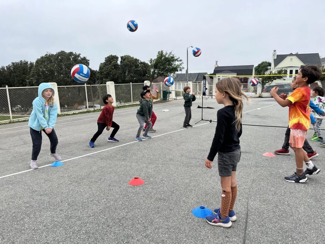Children tossing volleyballs into the air on an outdoor court.