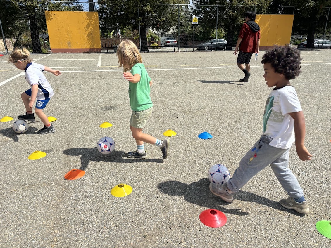 Children moving with soccer balls between cones on an outdoor court.