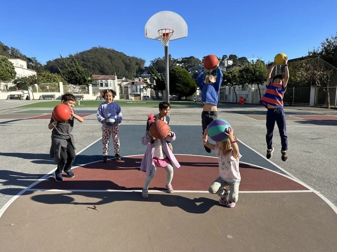 Children raising basketballs toward a hoop on an outdoor court.