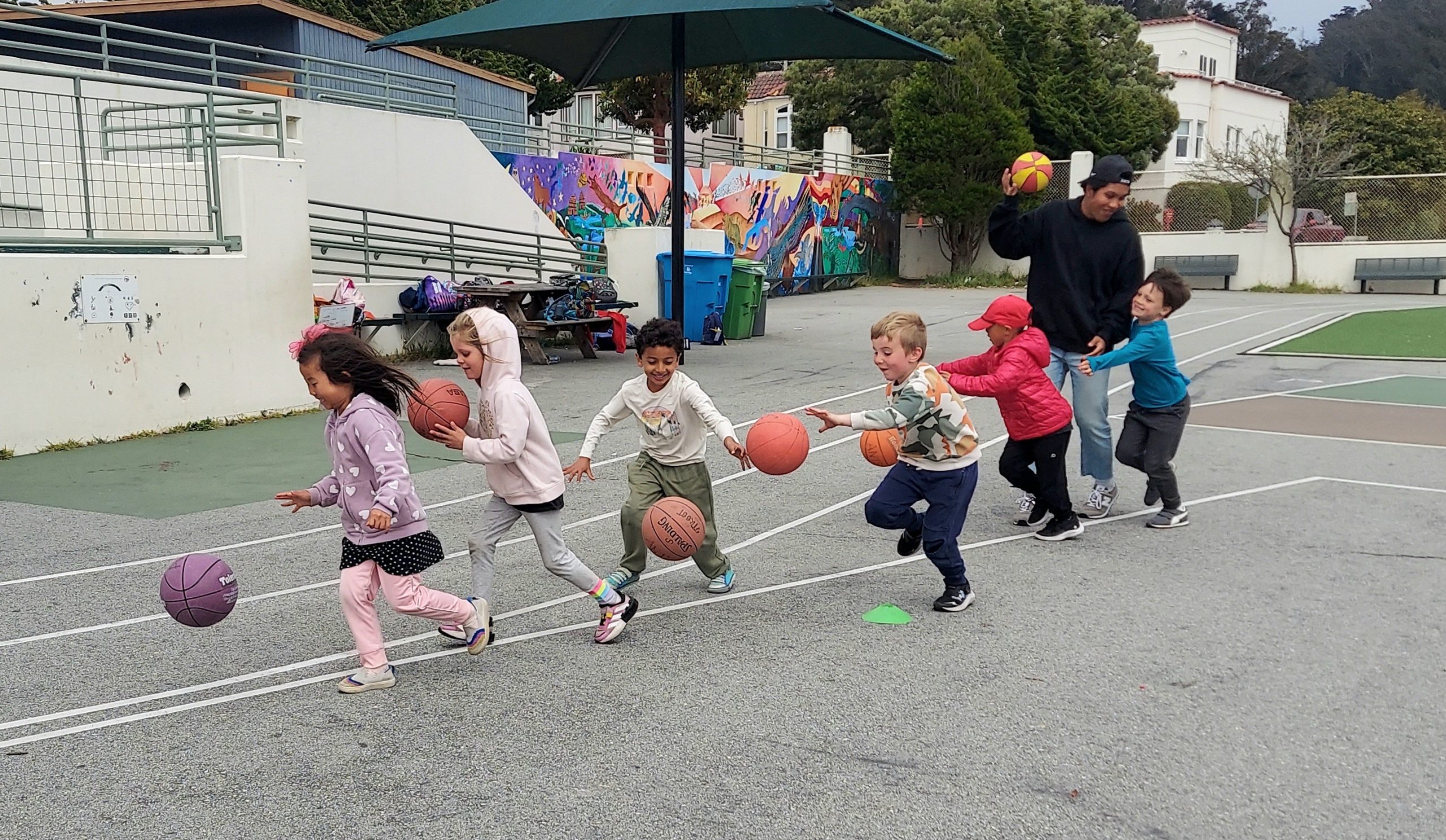 Children dribbling basketballs on an outdoor court with a coach.