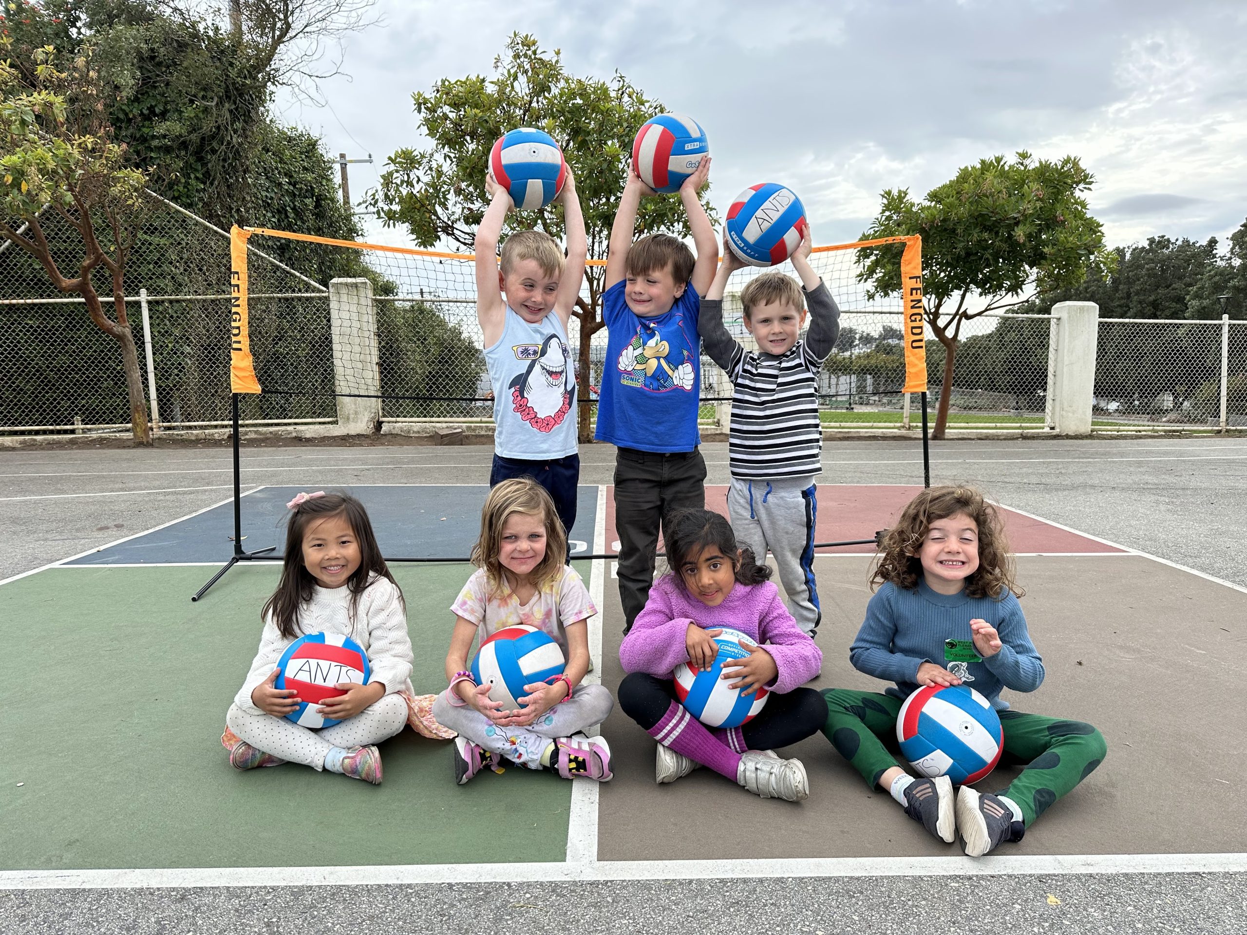 Children posing together with volleyballs beside an outdoor net.