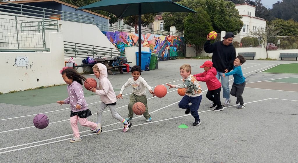 Children dribbling basketballs with a coach on a school playground.