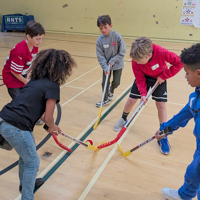 Children playing floor hockey with an ANTS coach in a gym.