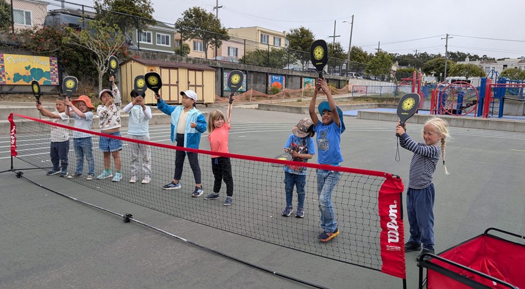 Children holding pickleball paddles behind a low net on a schoolyard.