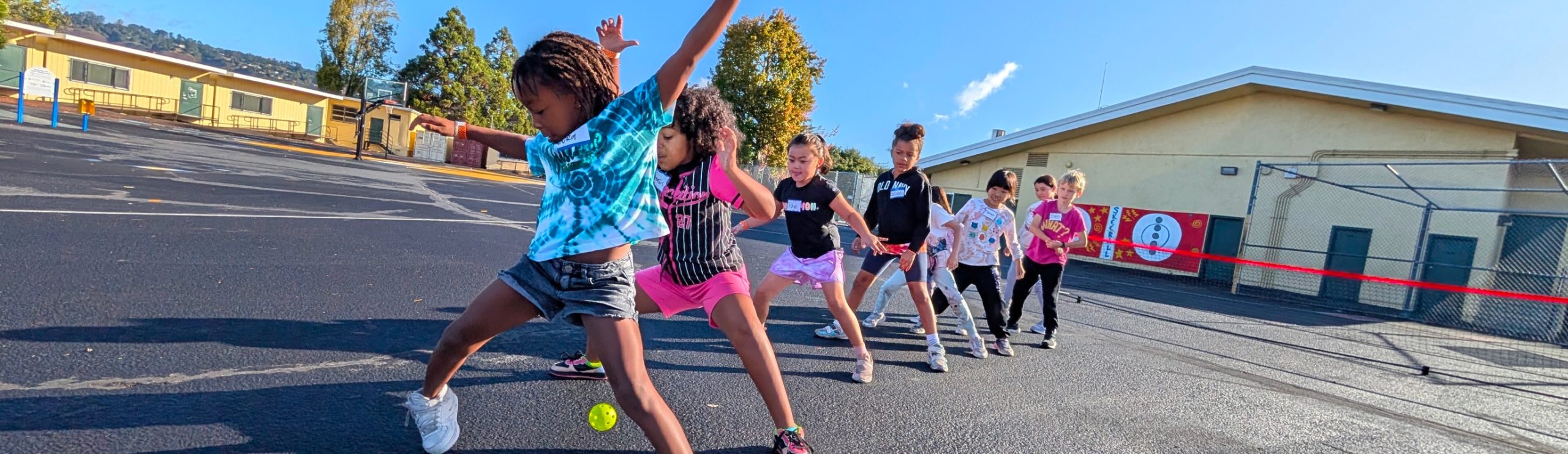Children playing tennis on a schoolyard with a ball near their feet.