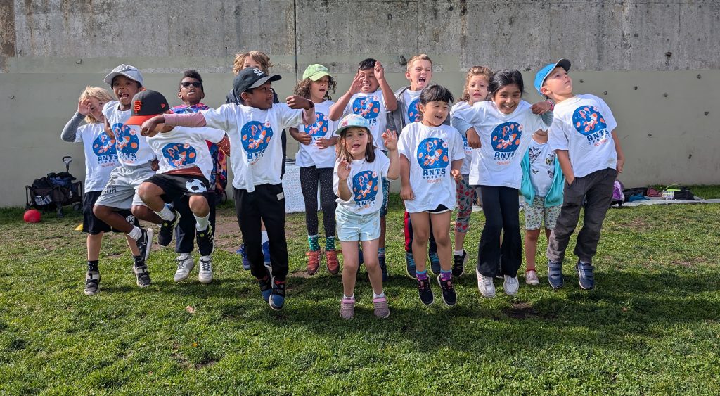 Children posing together in white ANTS camp shirts on the grass.