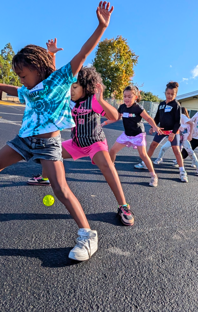 Children playing tennis on a schoolyard with a ball near their feet.