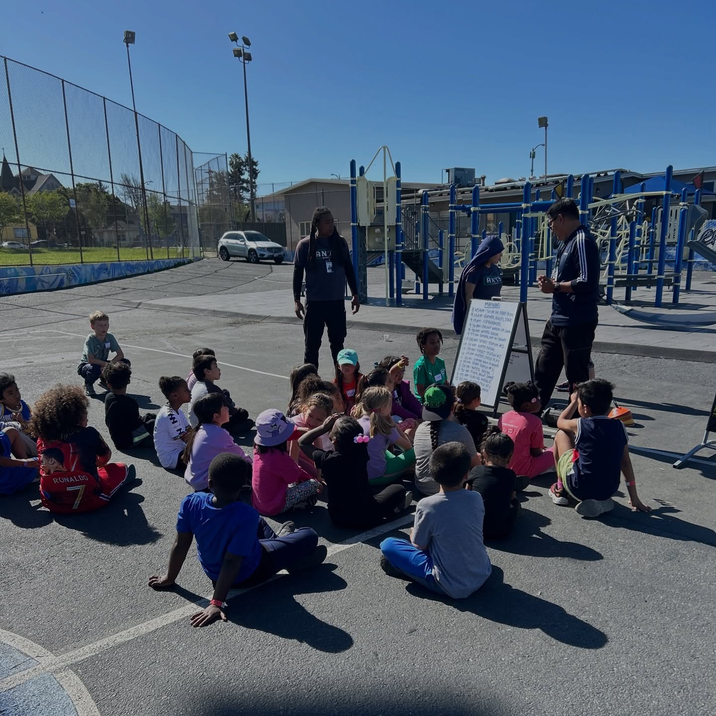Children seated on an outdoor court with ANTS coaches standing in front of them.