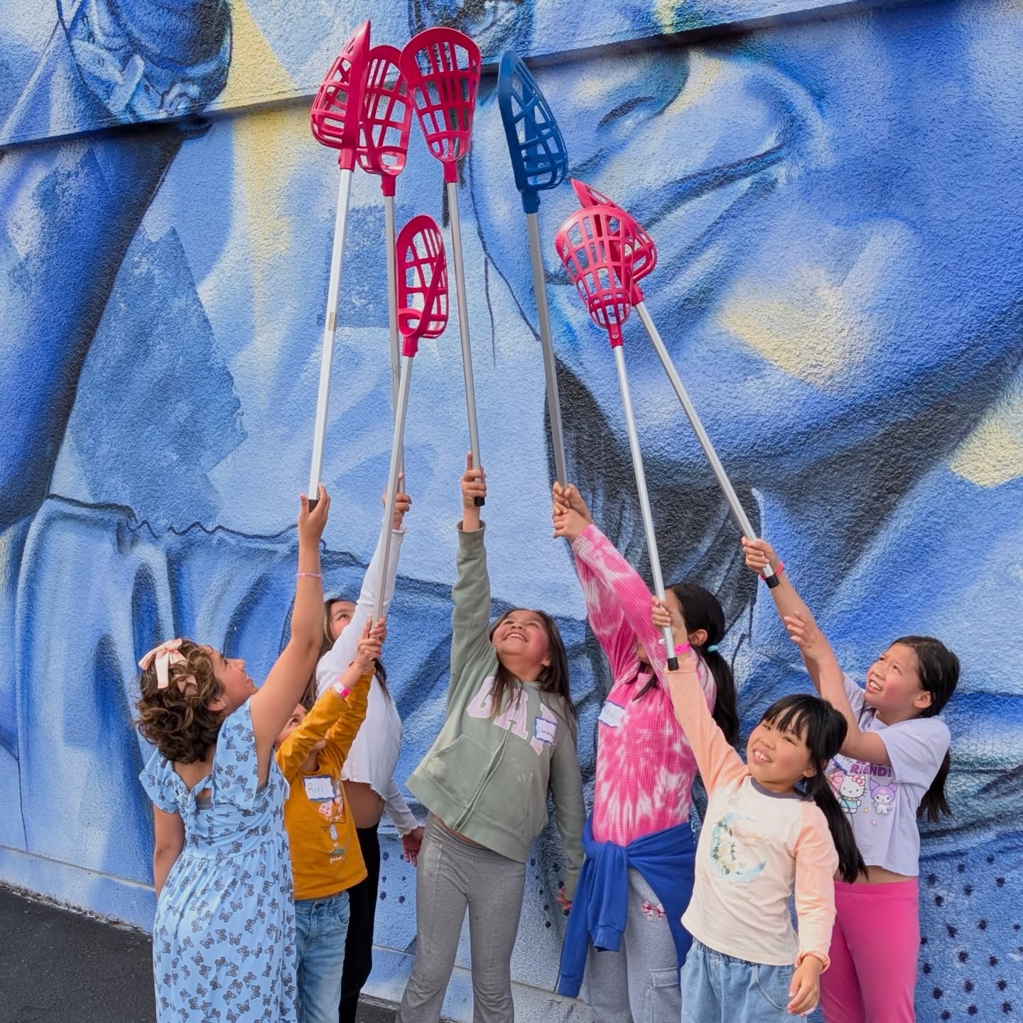 Children raising lacrosse sticks together in front of a mural.