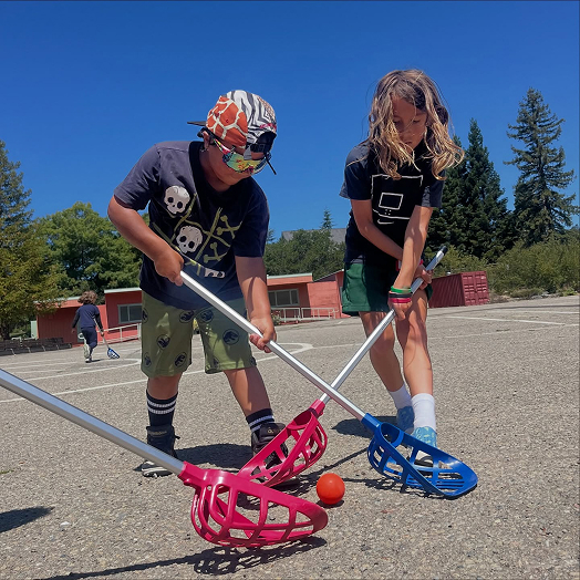 Two children using floor hockey sticks on an outdoor court.