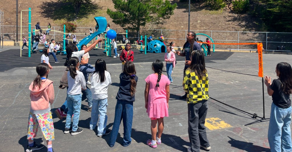 An ANTS coach leading a ball activity with children beside a playground.