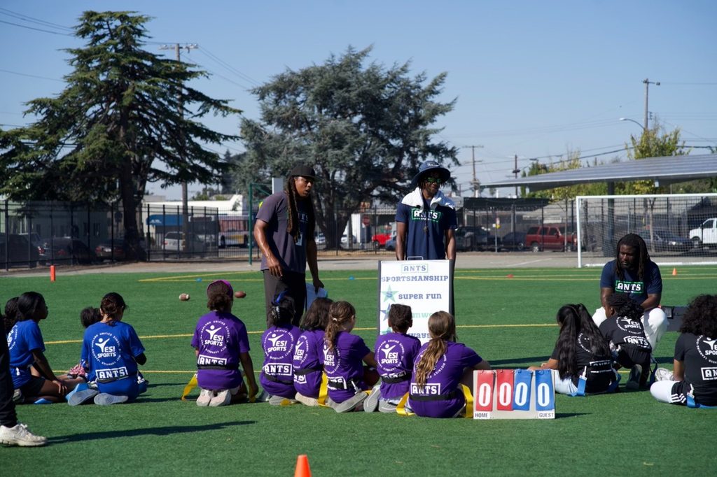 Children seated on a field beside a scoreboard with ANTS coaches.