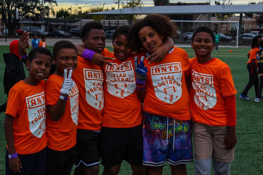 Children in orange ANTS flag football shirts posing together on a field.